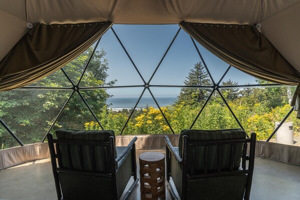 A room with a view of the ocean, featuring a table with two chairs and a wooden stool.