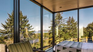 A bedroom with a large window, a bed, a chair, and a view of trees and a cliff. - South Cape Mirror Cabin, Queen - Floor to Ceiling Expansive Views (Cloverdale)