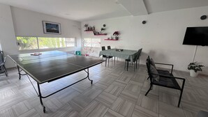 Une pièce spacieuse équipée d’une table de ping-pong, d’une table à manger et de chaises. De grandes fenêtres offrent une vue sur la verdure environnante.