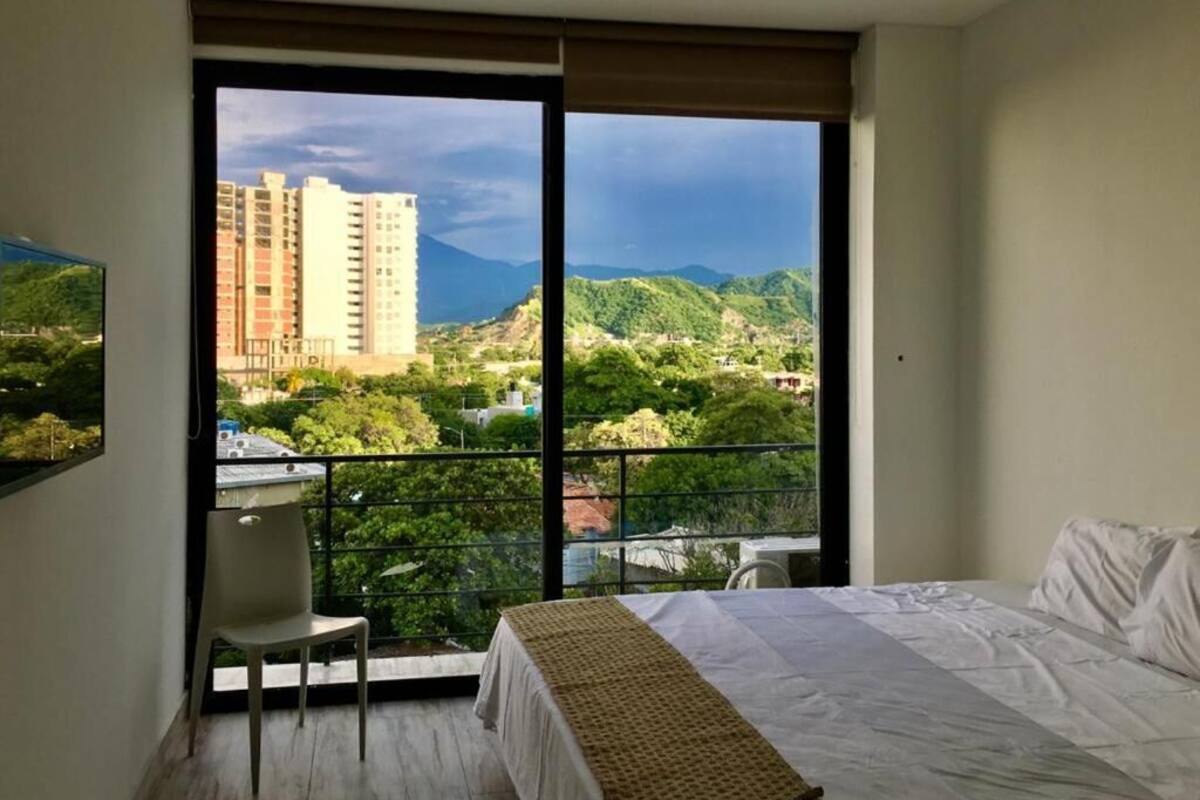 Un dormitorio con cama, una silla y un ventanal con vistas a edificios y zonas verdes.
