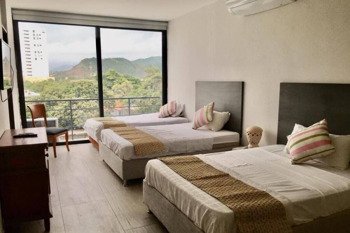 Habitación de hotel con dos camas, un escritorio y una silla. La habitación tiene un ventanal con vistas a zonas verdes y edificios.