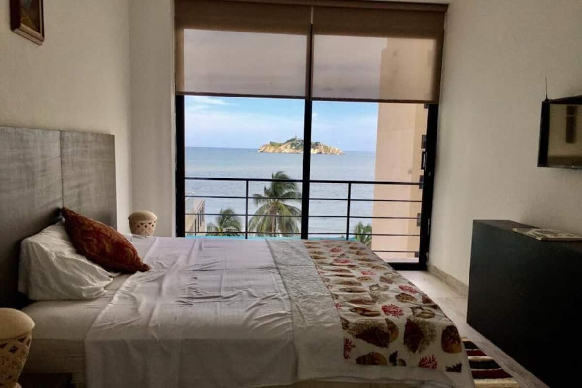 Un dormitorio con cama, una ventana que ofrece vista al mar y una pequeña isla, y un mueble oscuro a la derecha.