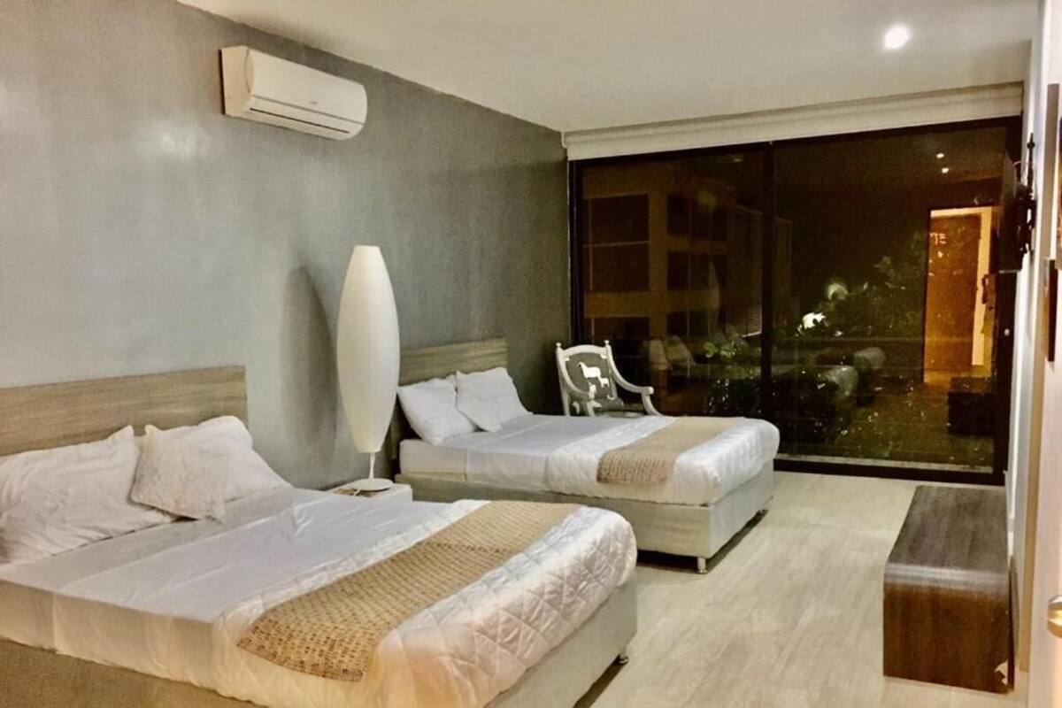 Habitación de hotel con dos camas, un ventanal grande, una silla y un equipo de aire acondicionado.