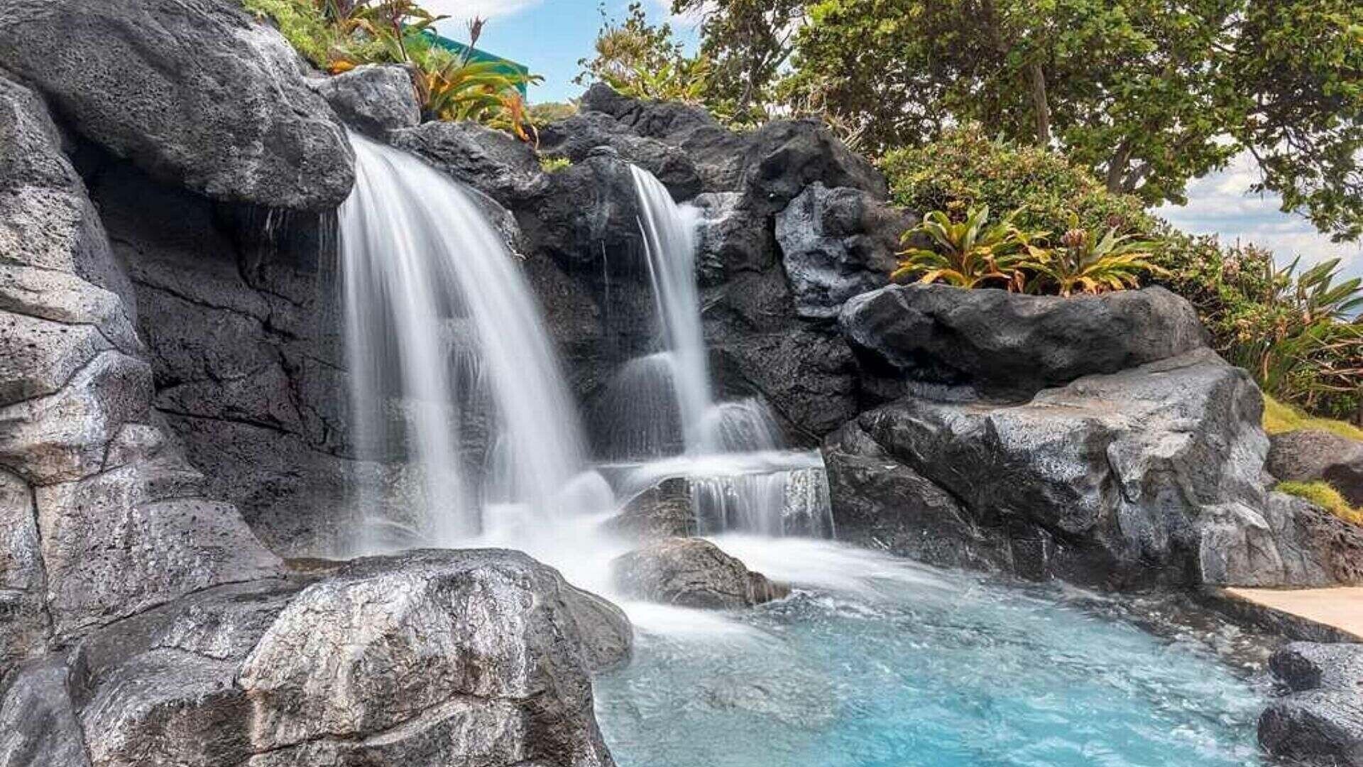 A waterfall cascading over dark rocks into a clear pool, surrounded by lush greenery.