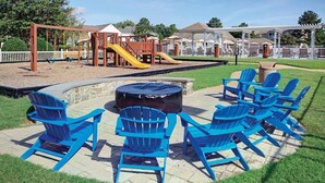 Ein Spielplatz mit Rutschen und Schaukeln, umgeben von einem weißen Zaun. Im Vordergrund stehen blaue Adirondack-Stühle und eine Feuerstelle aus Stein.
