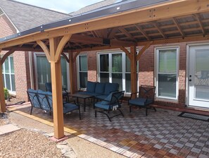 A covered patio area with outdoor furniture and a brick building in the background.