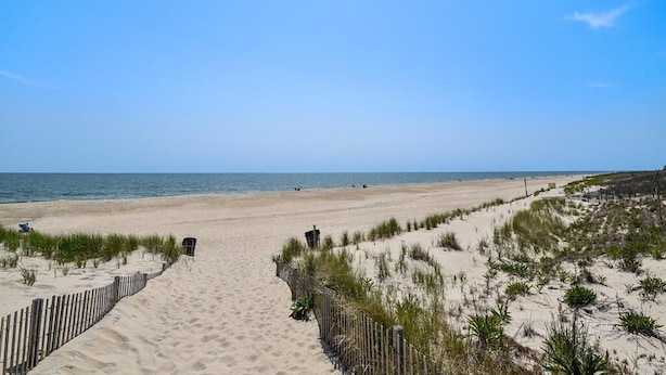 Ein Sandweg am Strand, der zum Meer führt, mit einer Holzlattenzaun und vereinzelter Vegetation.