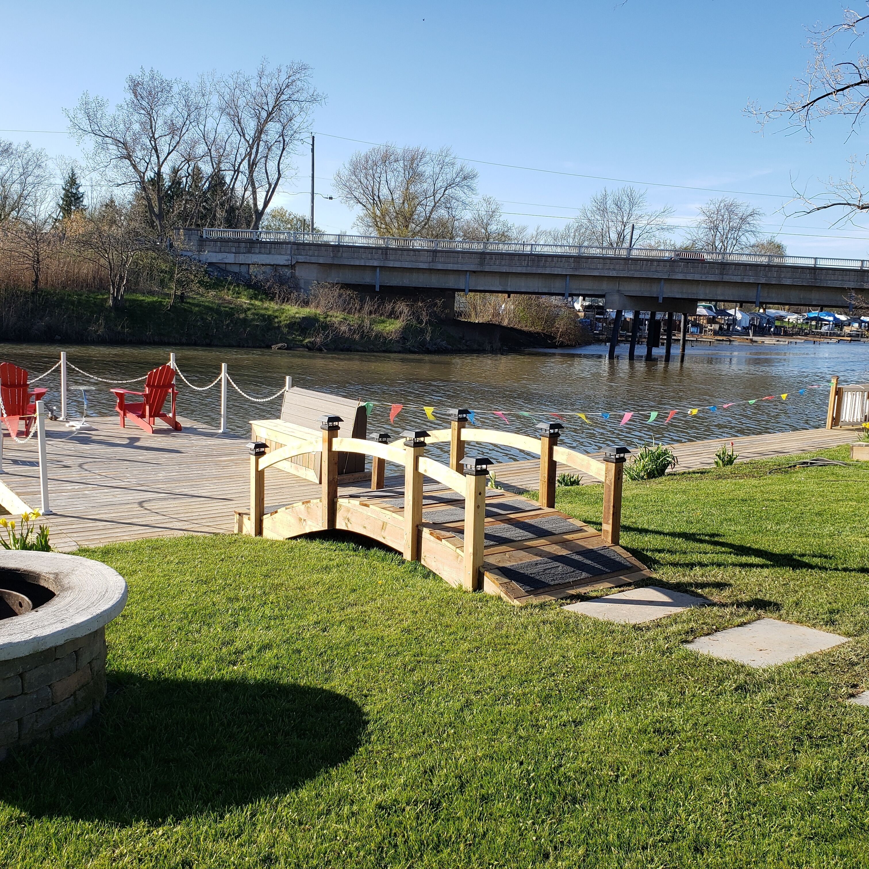Un parc au bord de la rivière, avec un pont en bois, des chaises rouges et une petite terrasse en bois.