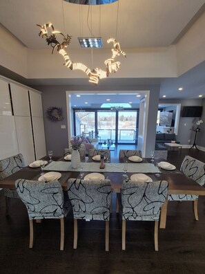 Un espace de repas moderne, avec une grande table dressée pour six personnes, entourée de chaises aux motifs originaux. Un luminaire unique est suspendu au plafond, et une vue sur l’extérieur est visible à travers de grandes fenêtres.