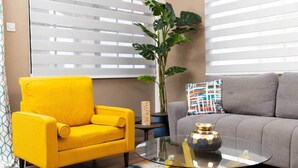 Un salon avec un fauteuil jaune, un canapé gris, une table basse en verre et une grande plante.