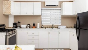 Une cuisine moderne avec des meubles blancs, un réfrigérateur noir, une cuisinière et une fenêtre équipée de stores.