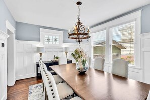 A dining area with a wooden table, white chairs, a chandelier, and a window with blinds. - Historic Home with King Bed (High Point)
