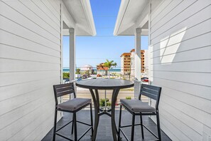 A covered outdoor seating area with a table and two chairs, overlooking a beach and buildings. - Sea Breeze Bliss Vacation Home (Fort Myers Beach)