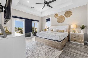 A modern bedroom with a large bed, a ceiling fan, a view of the outdoors, and a wooden nightstand with a lamp. - Sea Breeze Bliss Vacation Home (Fort Myers Beach)