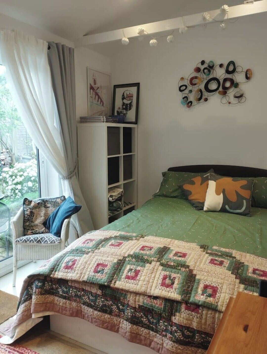 A bedroom with a green bedspread, a quilt, a chair with a patterned cushion, a shelf with books, and a wall art piece.