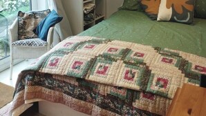 Une chambre avec une couvre-lit verte, une courtepointe, une chaise avec un coussin à motifs, une étagère remplie de livres et une œuvre d’art murale.