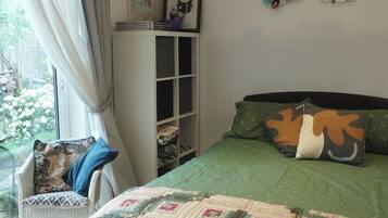 A bedroom with a green bedspread, a quilt, a chair with a patterned cushion, a shelf with books, and a wall art piece.