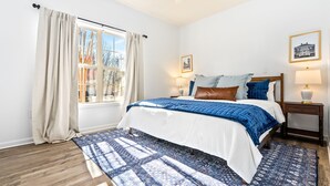 A bedroom with a bed, a window with curtains, a nightstand with a lamp, and a wall with a framed picture.