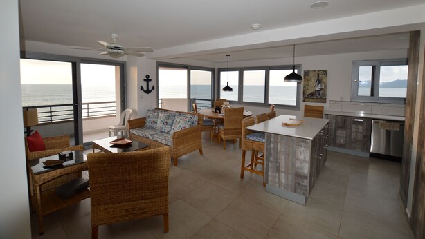 A spacious living area with a balcony overlooking the sea, wicker furniture, a dining table, and a kitchen with modern appliances.