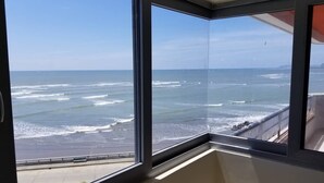 A room with a large window overlooking a beach with waves and a clear sky.