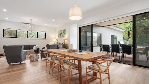 A spacious living area with a wooden dining table, comfortable seating, and large windows offering a view of the outdoors. - The Cape House in Cape Woolamai (Cape Woolamai)