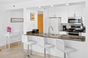 A modern kitchen with white cabinetry, a granite countertop, stainless steel appliances, and three white bar stools. - Brickell City Centre Condo with Pool & Parking (Miami)