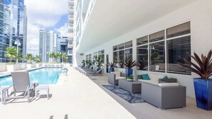 Un espace piscine aménagé sur le toit, avec des chaises longues et des canapés, entouré de grands immeubles.