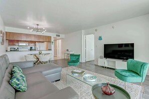 Ein modernes Wohnzimmer mit einem grauen Sofa, einem Glastisch und einem grünen Sessel. Im Hintergrund ist eine Küche mit Holzschränken und einer weißen Arbeitsplatte zu sehen.