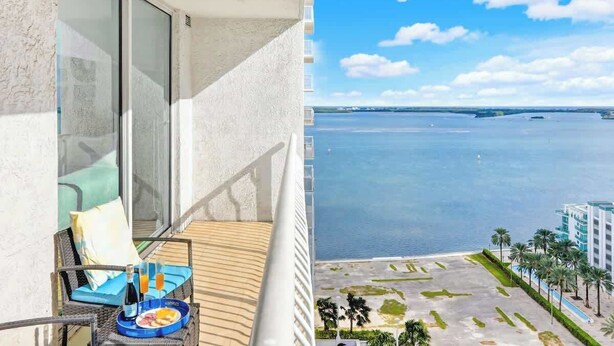 A balcony with a dining setup, a view of a calm body of water, and a distant shoreline with buildings. - Brickell Condo w Pool, Gym, Balcony & Parking (Miami)