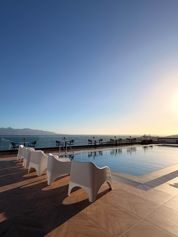 A rooftop pool with clear views of the sea and mountains, surrounded by white chairs and tables.
