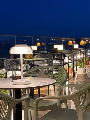 Terrasse mit runden Tischen und Stühlen, Beleuchtung und Blick auf die Stadt bei Nacht.