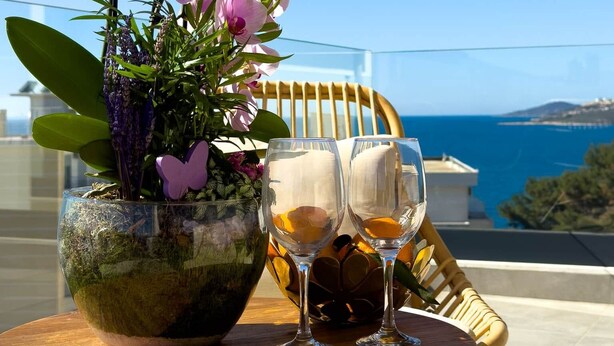 Een tafel met een vaas roze orchideeën en twee wijnglazen, op een balkon met uitzicht op zee.