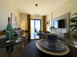 A modern hotel room with a dining area, a sofa, a TV mounted on the wall, and a view of the city. - Dori Coastal Escape Saranda (Sarandë)