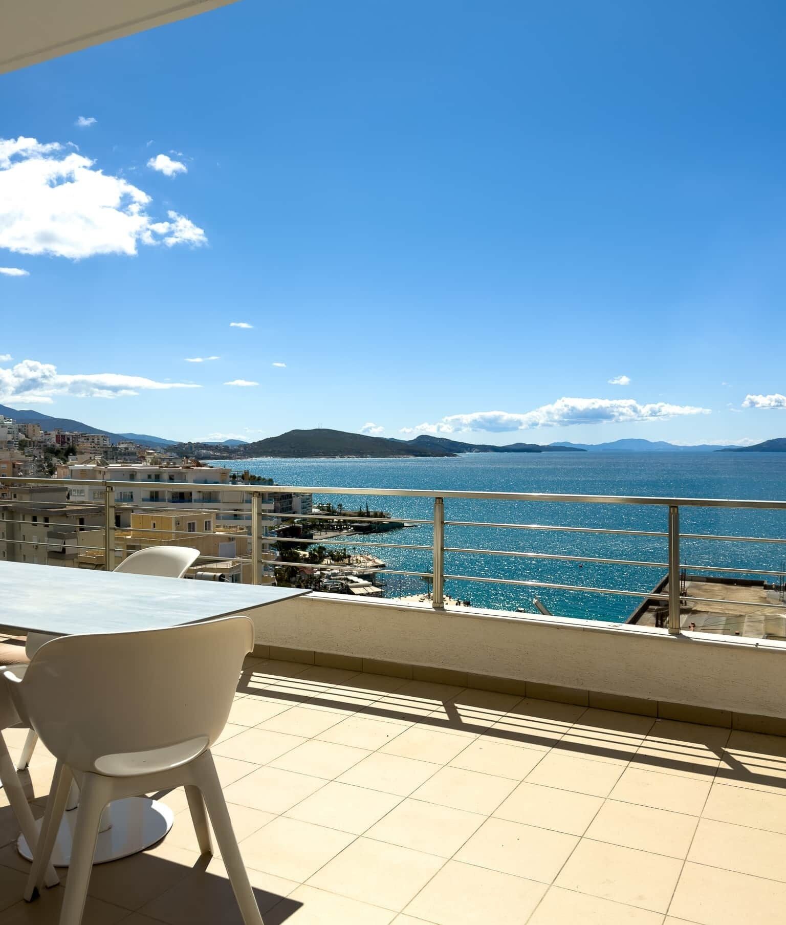 A terrace with a table and chairs, overlooking a coastal view with buildings and mountains in the distance.