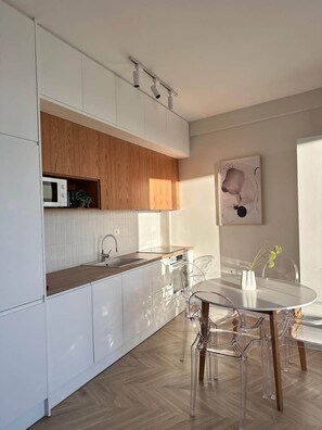A modern kitchen with white cabinets, a wooden upper section, a microwave, and a dining area with a glass table and chairs. - Valentina - Corfu View Residence (Sarandë)