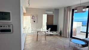 A modern living room with a herringbone floor, a dining area with clear chairs, a flat-screen TV, and a large window offering a view of the sea. - Valentina - Corfu View Residence (Sarandë)