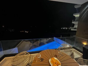 A balcony with a table and chairs, a glass partition, and a view of the city at night. - Veronica - Corfu View Residence (Sarandë)