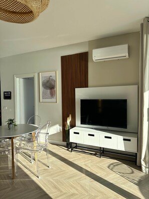 A modern living room with a large flat-screen TV, a dining area with a round table and clear chairs, a potted plant, and a wall-mounted air conditioner. - Veronica - Corfu View Residence (Sarandë)
