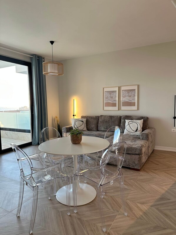 Een moderne woonkamer met een ronde witte tafel, transparante stoelen, een grijze bank en twee ingelijste foto's aan de muur.