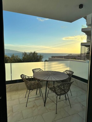 Een balkon met een ronde tafel en rieten stoelen, met uitzicht op een waterpartij en verre bergen.
