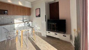 Un salon moderne comprenant un téléviseur, une table à manger avec des chaises, et un coin cuisine avec des placards et des appareils électroménagers.