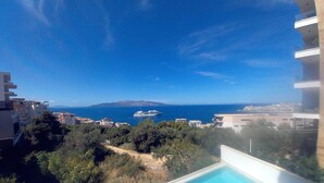 Une vue depuis la piscine donnant sur une zone côtière, avec des bâtiments et un navire de croisière à l’horizon.