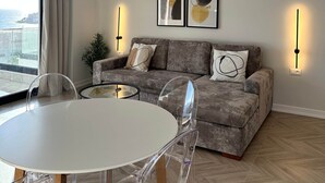 En moderne stue med en grå sofa, et rundt spisebord med klare stole og et stort vindue med gardiner.