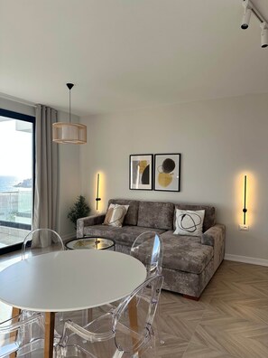 A modern living room with a grey sofa, a round dining table with clear chairs, and a large window with curtains. - Victoria - Corfu View Residence (Sarandë)