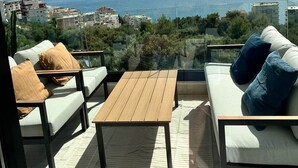 Un balcon avec un canapé, une table en bois et une vue sur la mer et les montagnes lointaines.