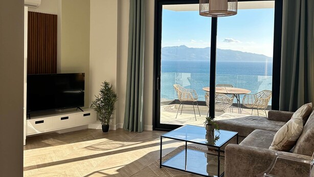 A modern living room with a large flat-screen TV, a sofa, a coffee table, and a view of the sea through large windows. - Vivien - Corfu View Residence (Sarandë)