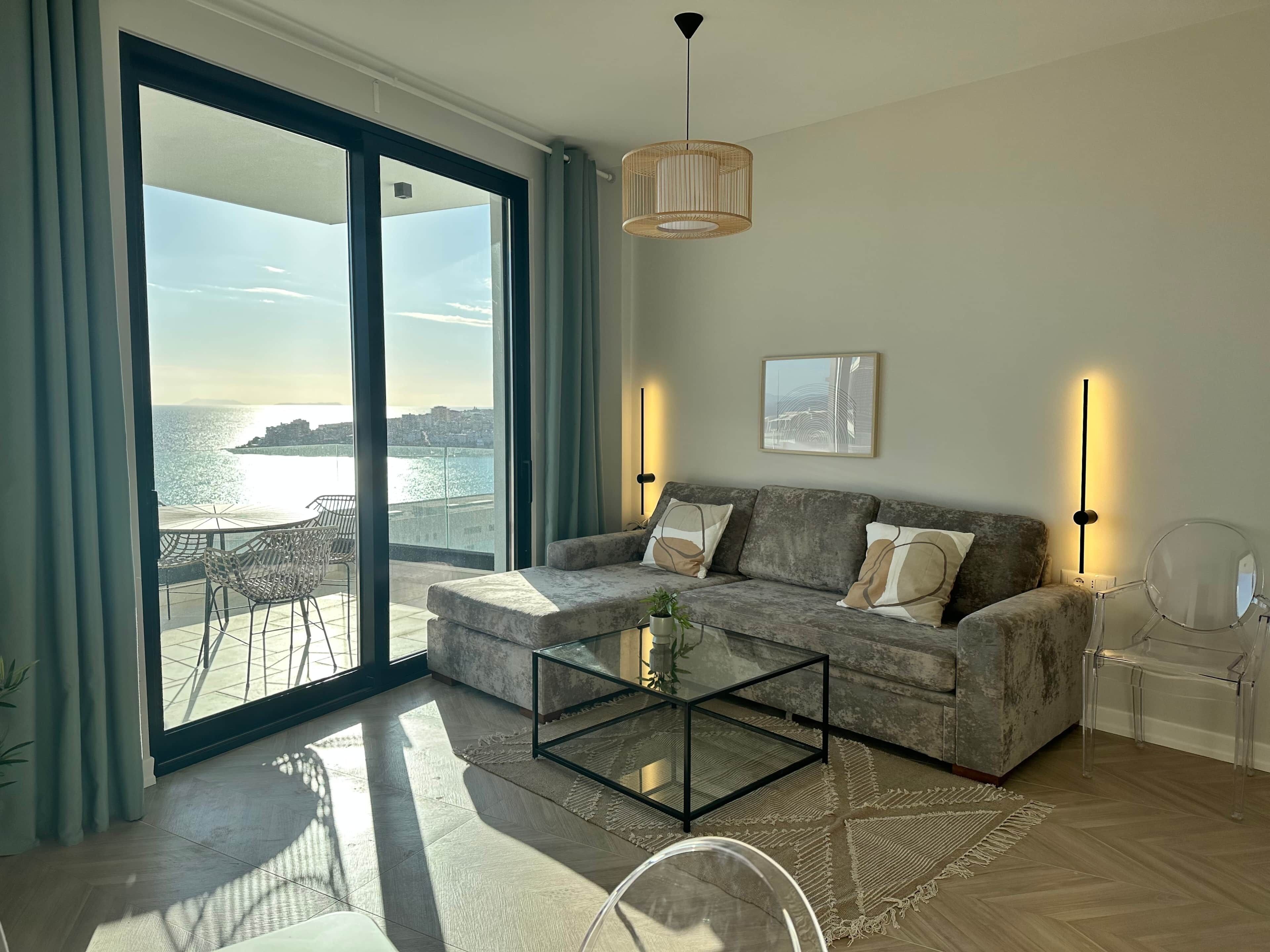 A modern living room with a grey sofa, a glass coffee table, and a view of the sea through large sliding doors.