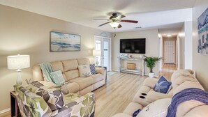 Un salon avec un canapé beige, un fauteuil à motifs, un ventilateur de plafond, un téléviseur à écran plat et une plante en pot.