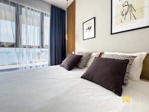 Habitación de hotel con una cama grande, dos almohadas, una obra de arte enmarcada en la pared y vista al mar a través de cortinas translúcidas.
