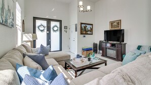 Un salon avec un canapé, une table basse et un meuble pour la télévision. On y trouve une porte décorée d’une guirlande, un tableau accroché au mur et un panier posé sur le sol.